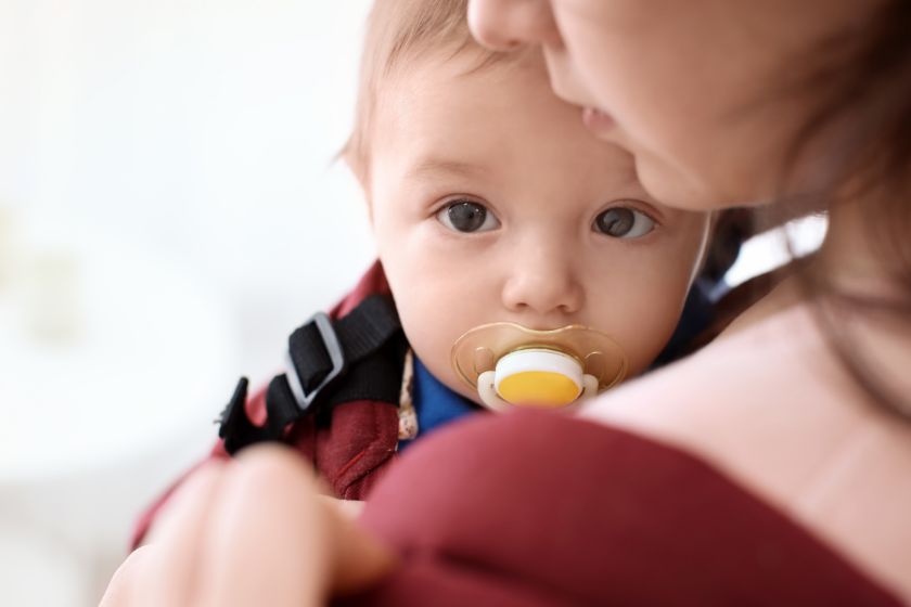 Ein Baby mit Schnuller schaut ruhig in die Kamera, während es von einer erwachsenen Person im Arm gehalten wird.