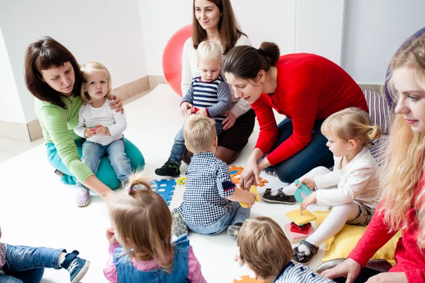 Vier Erzieherinnen und 6 kleine Kinder sitzen zusammen am Boden. Um sie herum liegt Spielzeug, mit welchem sie gerade spielen und zwei der Kinder sitzt auf dem Schoß zwei Erzieherinnen.