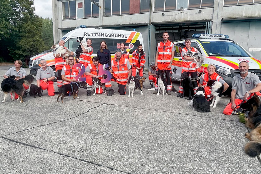 Mehrere Johanniter stehen zusammen mit einigen Hunden vor Johanniter Einsatzfahrzeugen und halten eine große 25 in der Hand.