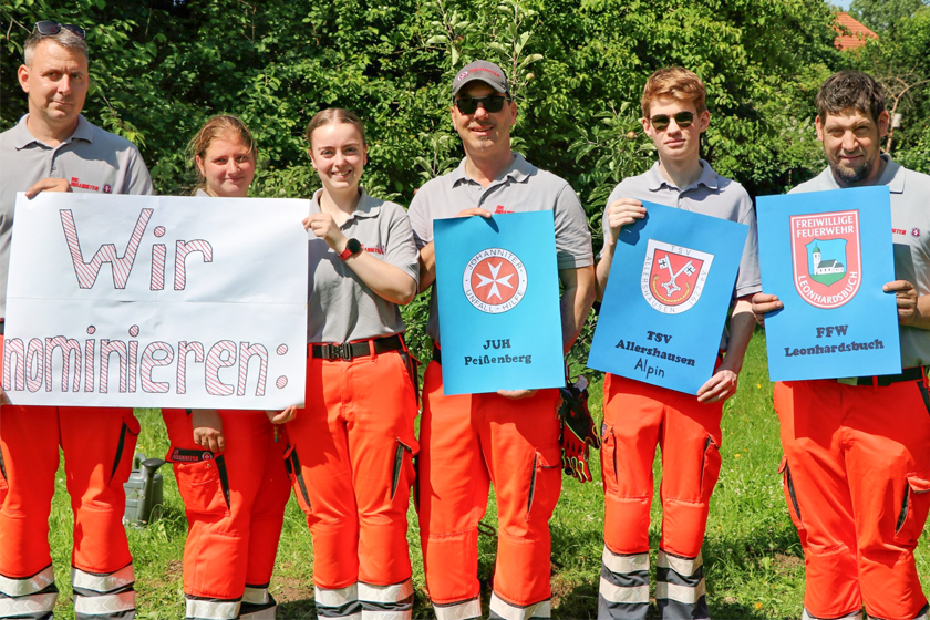 Mehrere Johanniter stehen vor einem Wald und halten folgende Schilder hoch: Wir nominieren: JUH Peißenberg, TSV Allershausen Alpin, FFW Leonhardsbuch.