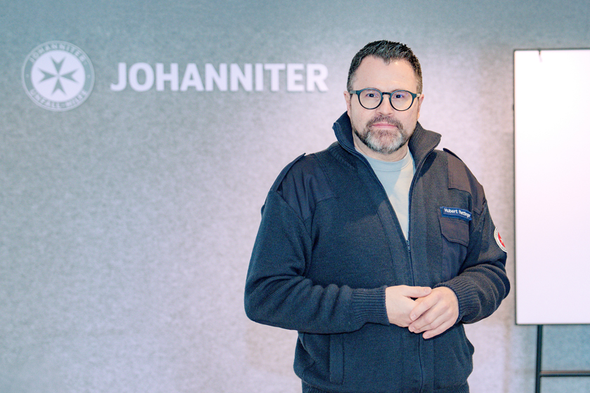 Ein Johanniter steht vor einer grauen Wand mit einem Johanniter Logo und schaut in die Kamera.