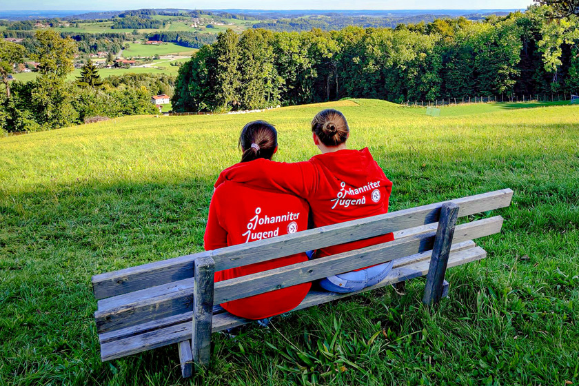 Zwei Frauen mit „Johanniter Jugend“ Pullovern sitzen auf einer Bank und blicken auf die Landschaft.