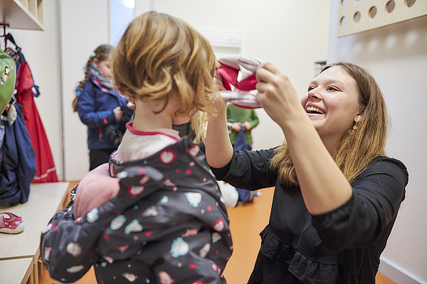 Eine Erzieherin hilft einem Kind im Kindergarten beim anziehen der Mütze