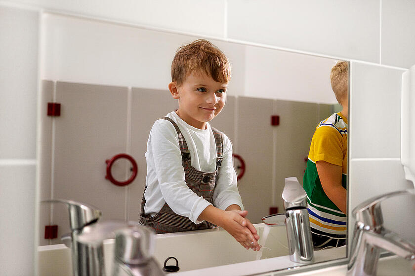 Ein kleiner Junge lächelt in den Spiegel und wäscht sich die Hände.