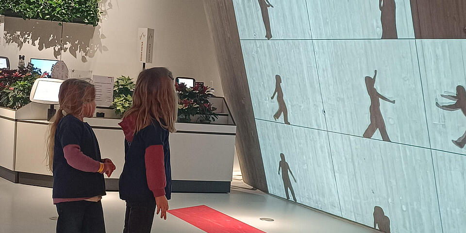 Zwei Mädchen schauen auf eine Betonwand, an der Schatten zu sehen sind.