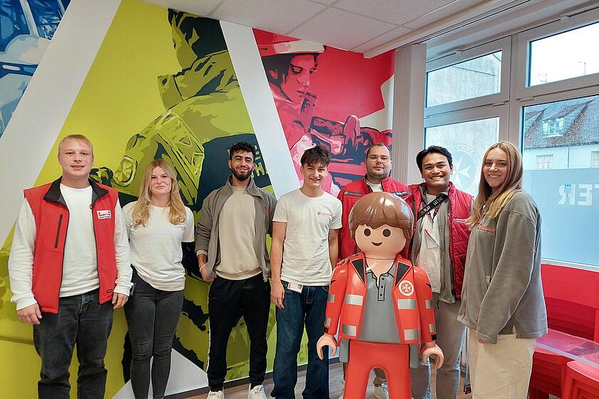 Eine Gruppe junger Leute in Johanniter Kleidung stehen um ein großes Johanniter Playmobilmännchen und lächeln in die Kamera.