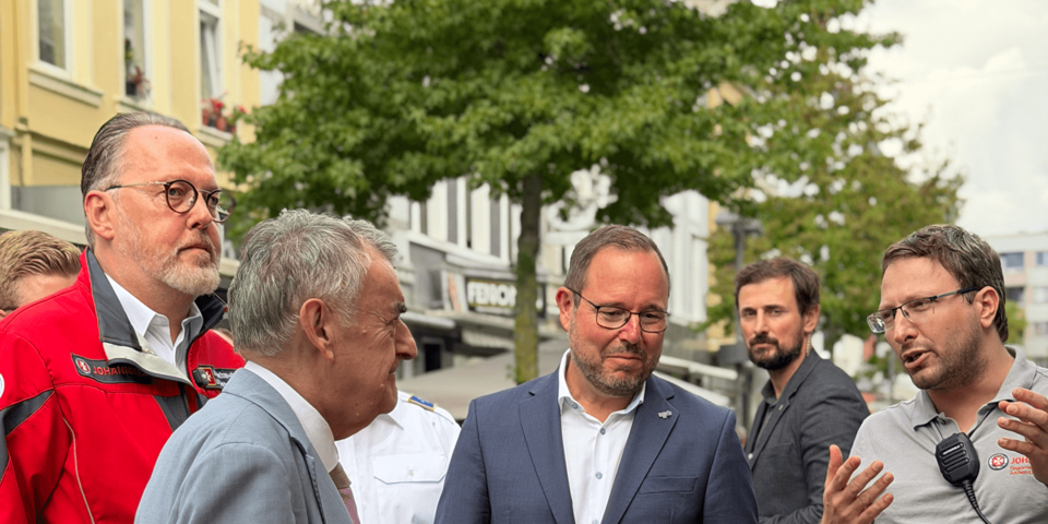 Männer in Geschäftsanzügen und in roter Johanniter-Einsatzkleidung stehen im Gespräch auf Straße.