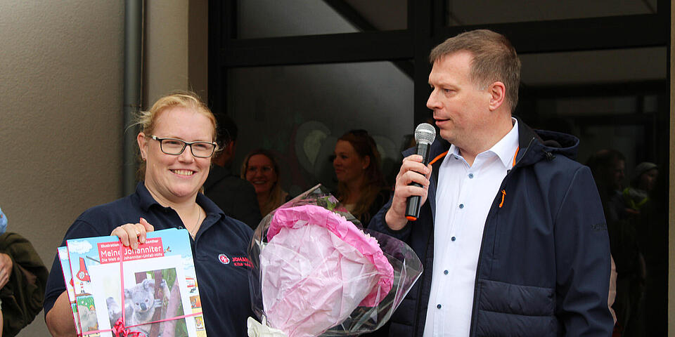 Links ein Mann mit Mikro. Daneben eine Frau mit Blumenstrauß und einem Kinderbuch.