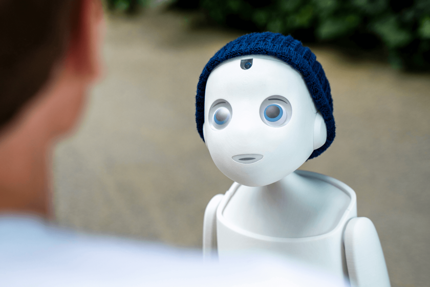 eine kleine weiße Figur mit großen Augen und blauer Mütze scheint einen Menschen zu betrachten, der ihm gegenübersteht und von dem nur ein Ausschnitt von Hals und Ohr sowie Schulter von hinten zu sehen sind.