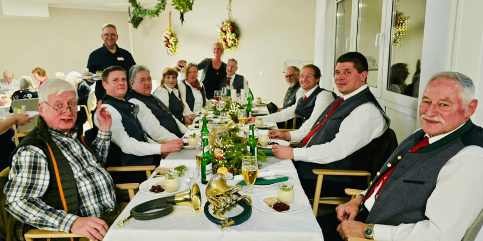 Lange festliche Tafel mit Jagdgesellschaft in Tracht, Menü und Getränken, grün geschmückt und weihnachtlich dekoriert.
