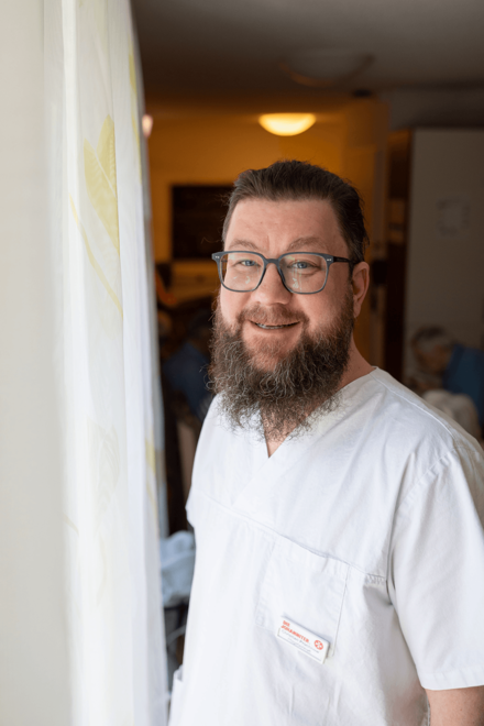 Ein Mann in weißer Arbeitskleidung steht am Fenster neben einem Vorhang, lächelt 