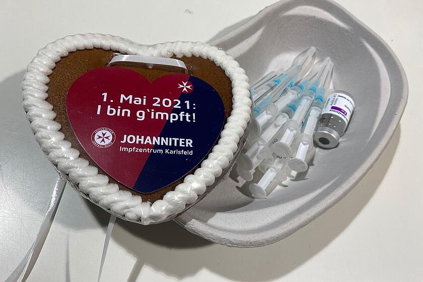 Ein Johanniter Lebkuchenherz mit der Aufschrift "01. Mai 2021 I bin g'impft!" liegt in einer Schüssel mit Impfstoff.