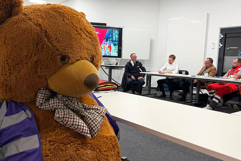 Im Vordergrund ist das Maskottchen, der PSNV-Teddy zu sehen und im Hintergrund Herr Kretschmer im Austausch mit den Ehrenamtlichen