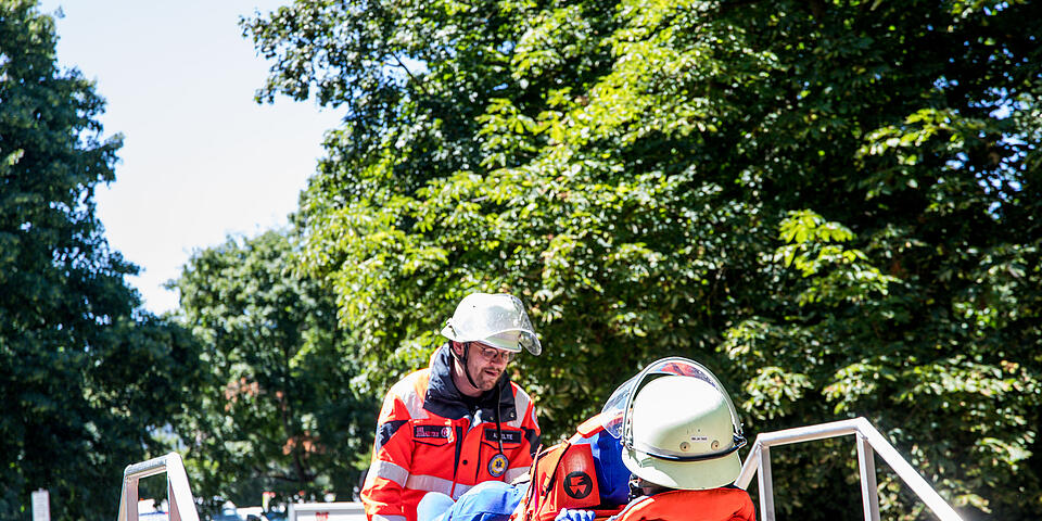 Rettungshelfer*innen in Schutzkleidung tragen auf einer Trage eine verletzte Person und arbeiten konzentriert bei einer Übung im Freien.