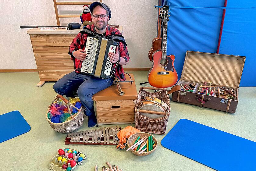 Musikpädagoge Lukas Johannsen hat ein Akkordeon in der Hand und sitzt vor einer großen Auswahl von verschiedenen Instrumenten im Bewegungsraum der Kita.