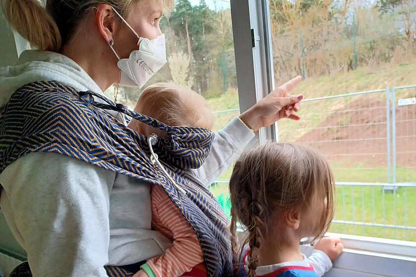 Erzieherin mit Kleinkind im Tragetuch und weiteres Kind neben sich am Fenster der Naturkita Schlüsselau
