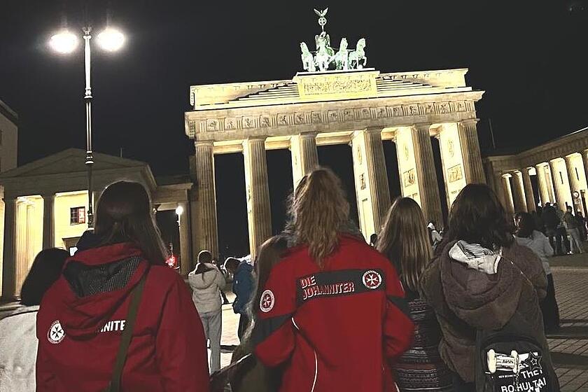 Das Foto zeigt die Reisegruppe der Ferienfreizeit, bestehend aus Kindern und Jugendlichen sowie den Betreuenden der Johanniter-Hochwasserhilfe. Die Gruppe läuft auf das Brandenburger Tor in Berlin zu, das angesichts der abendlichen Dunkelheit hell erleuchtet ist.