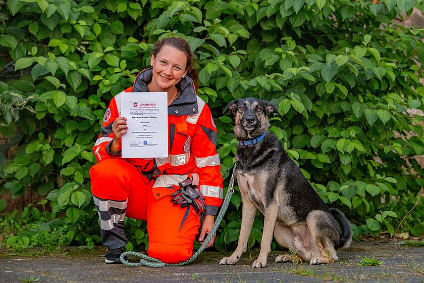 Eine junge Frau kniet neben ihrem Hund und hält ein Zertifikat hoch