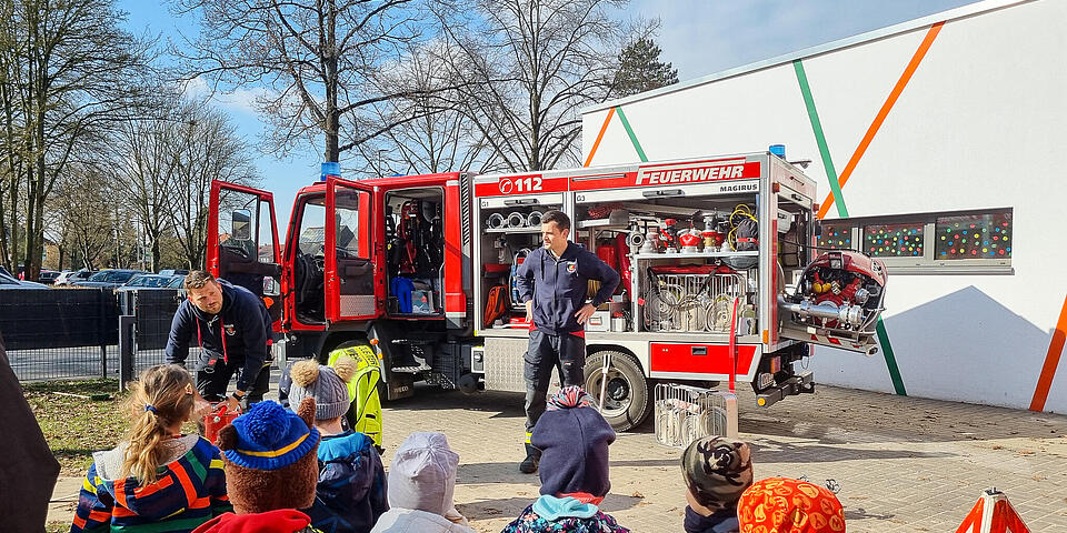Eine Gruppe von Kindern schauen auf ein Feuerwehrauto. Davor steht ein Mann in Feuerwehrbekleidung