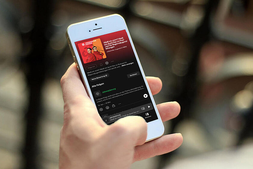 Ein Smartphone in der Hand einer Person. Auf dem Display ist ein Bild von zwei jungen Erwachsenen in Johanniter-Jugend-Begleidung zu sehen, die Rücken an Rücken zueinander stehen. Dies ist das Titelbild des Podcasts.