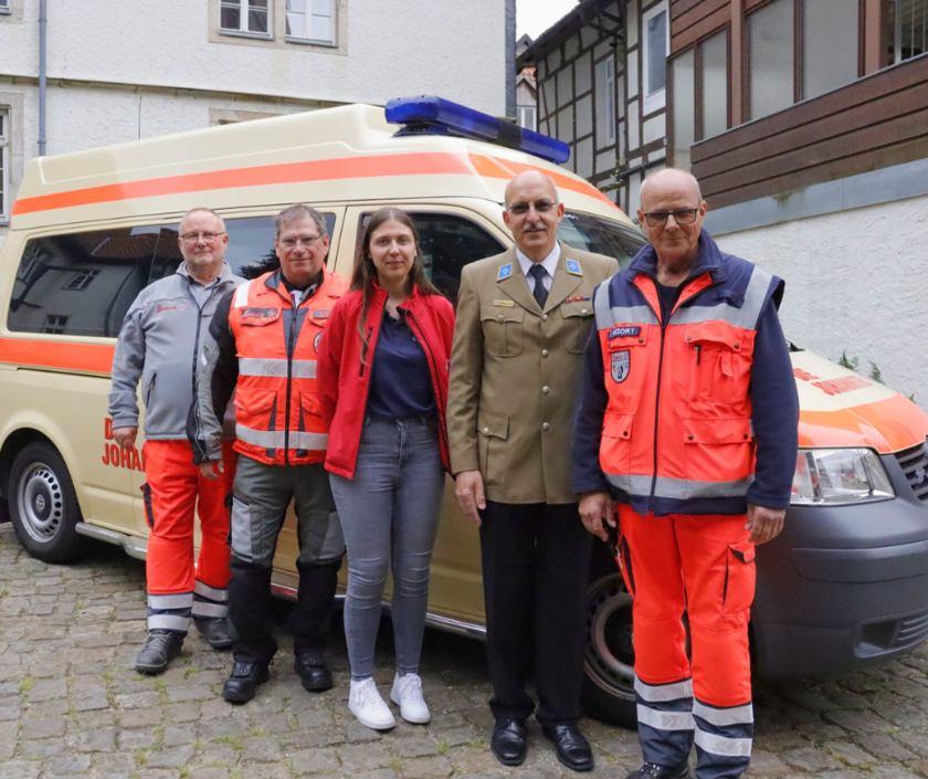 Fünf Ehrenamtliche der Johanniter stehen vor einem Einsatzfahrzeug. Alle tragen verschiedene Johanniter-Einsatz- oder Festkleidung und lächeln.