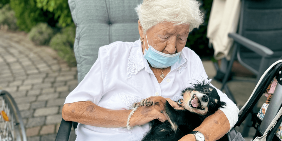 Eine weißhaarige Frau sitzt in einem Gartenstuhl. Sie trägt eine weiße Bluse und streichelt einen kleinen Hund.