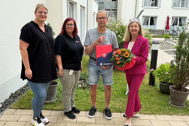 Drei Frauen und ein Mann stehen auf einem Gruppenbild mit Überreichung eines Blumenstraußes