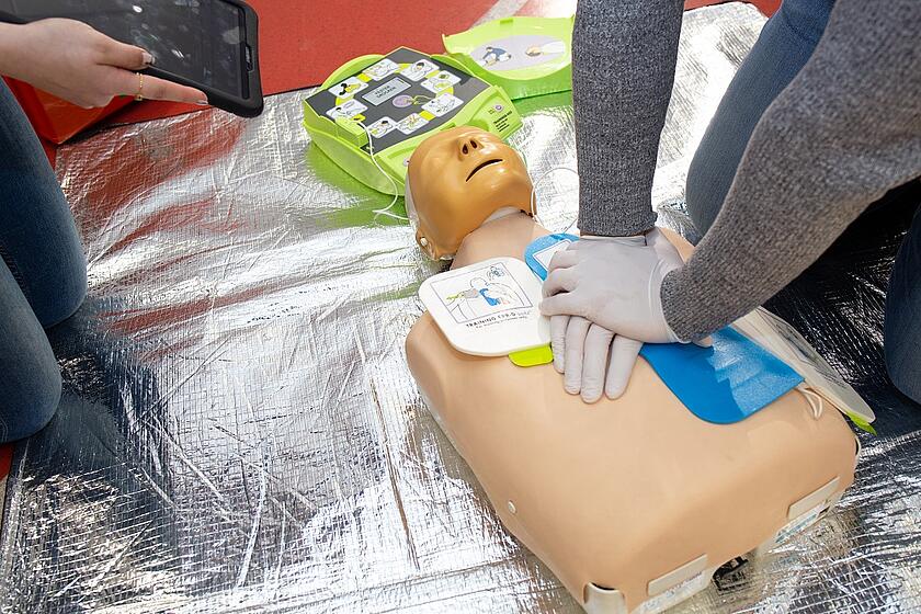 Zu sehen ist der Oberkörper und Kopf einer Puppe. Zwei Arme stützen sich mit übereinanderliegenden Händen auf den Herzbereich der Puppe. Die Person übt offensichtlich die Herz-Lungen-Wiederbelebung.