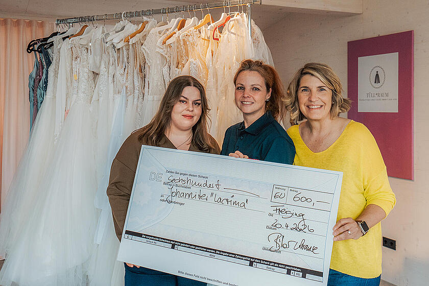 4 Frauen stehen vor aufgehangenen Hochzeitskleidern und halten einen Spendencheck für Lacrima.