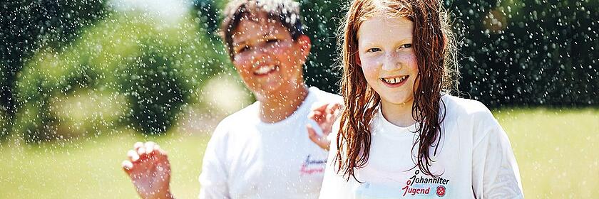 Zwei Kinder mit nassen Haaren und weißen T-Shirts mit „Johanniter Jugend“-Logo spielen fröhlich im Wasser.