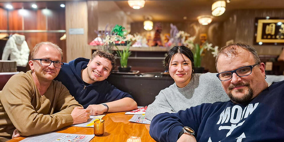 Ein junger Mann, zwei weitere Männer und eine junge Frau sitzen in einem Restaurant an einem Tisch. 
