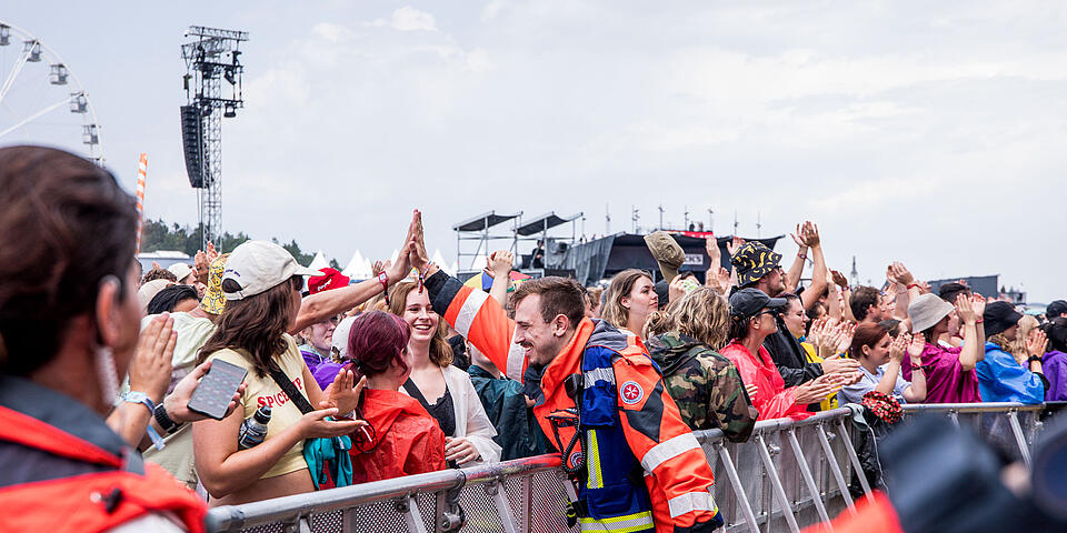Johanniter-Rettungssanitäter in Einsatzkleidung gibt Festivalbesucher*in an der Bühnenabsperrung ein High‑Five
