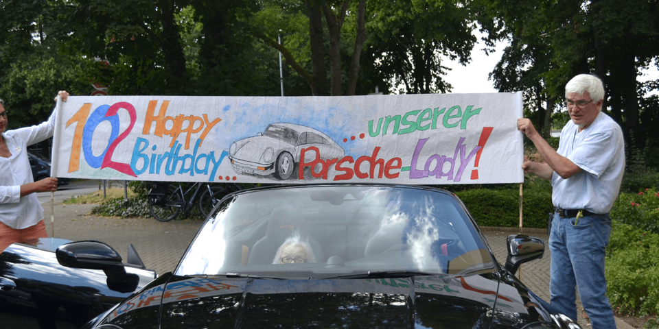 Eine Frau und ein Mann halten über das Dach eines Porsches ein Banner mit der Aufschrift "102 Happy Birthday unserer Porsche-Lady"