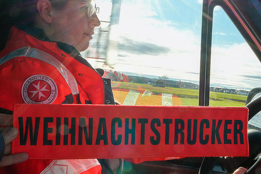 Eine Johanniterin in Einsatzkleidung fährt ein Auto und eine Hand hält ein Weihnachtstrucker Schild.