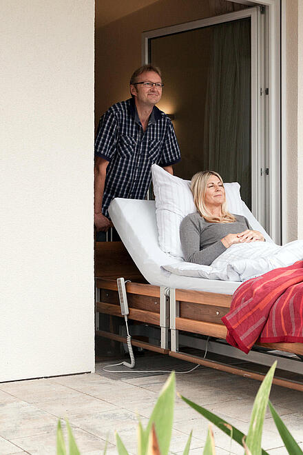 Frau im Pflegebett wird von ihrem Mann auf die Terrasse geschoben