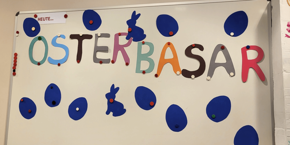 Auf einer weißen Tafel steht in bunten Buchstaben "Osterbasar". Darum blaue Osterhasen und Eier