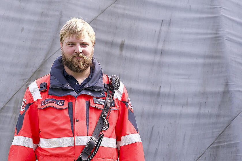Ein Mann mit blondem Haar und Bart trägt eine rote Einsatzjacke mit reflektierenden Streifen und dem Johanniter-Logo, dazu ein Funkgerät über der Schulter.
