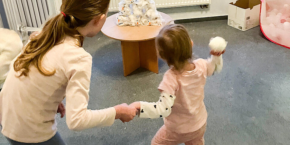 Ein kleines Kind wird von einem älteren Mädchen an der Hand gehalten und wirft einen Wattebaumball auf einen Schneemann aus Pappe