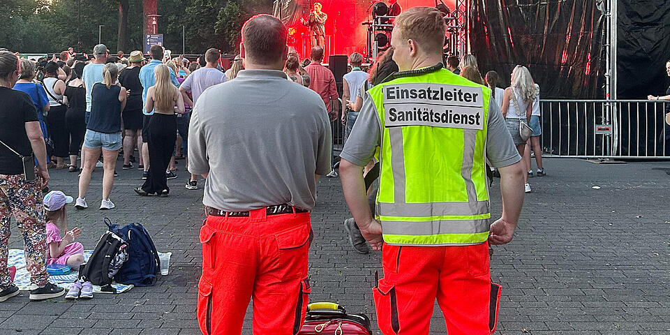 Zwei Männer in Einsatzkleidung mit einem Notfallrucksack zwischen ihnen vor einer Bühne auf der eine Band spielt. Einer trägt eine gelbe Weste 