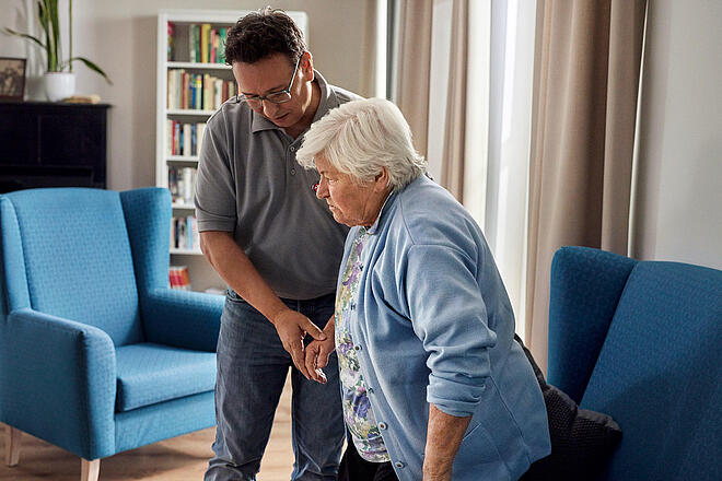 Pflegekurs für Angehörige Ein Mitarbeiter der Johanniter stützt eine Seniorin beim Aufstehen aus einem Sessel.