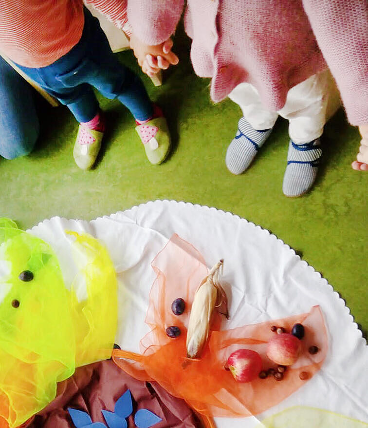 Eine Kindergartengruppe steht im Kreis um bunte Tücher auf denen Essen liegt um das Erntedankfest zu feiern.
