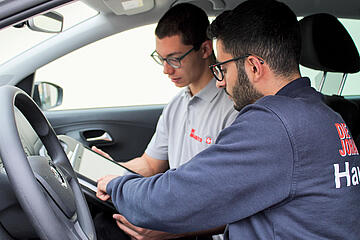 Zwei Männer im Auto; einer zeigt dem anderen Informationen auf einem Tablet.