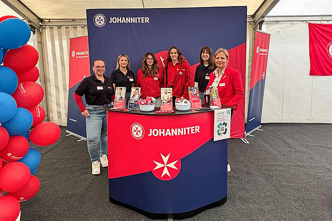Sechs Frauen mit Johanniter-Oberbekleidung stehen hinter einem Messepult. Hinter ihnen ist eine Stellwand. Alle Gegenstände haben ein Johanniter-Branding.
