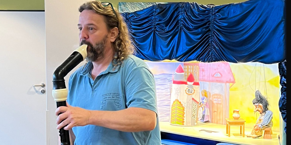 Ein Mann in einem blauen Poloshirt, schulterlange lockige Haar und Bart. Er hat eine Brille in die Haare gesteckt und spielt auf eine schwarzen Bassflöte. . Er steht vor einem Kasten mit blauem Vorhang, der der den Blick auf eine erleuchtete kleine Bühne freigibt.