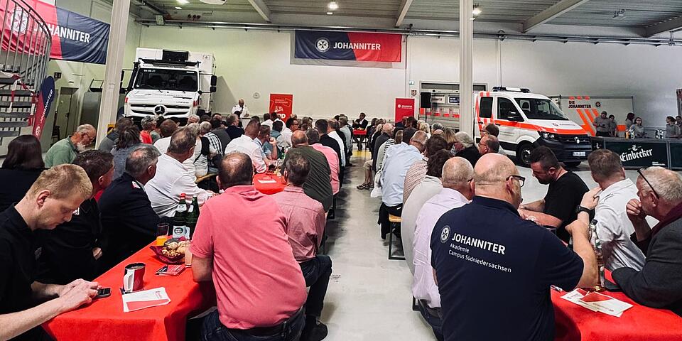 Menschen sitzen an Bierzeltgarnituren in einer großen Fahrzeughalle und schauen auf ein Podium