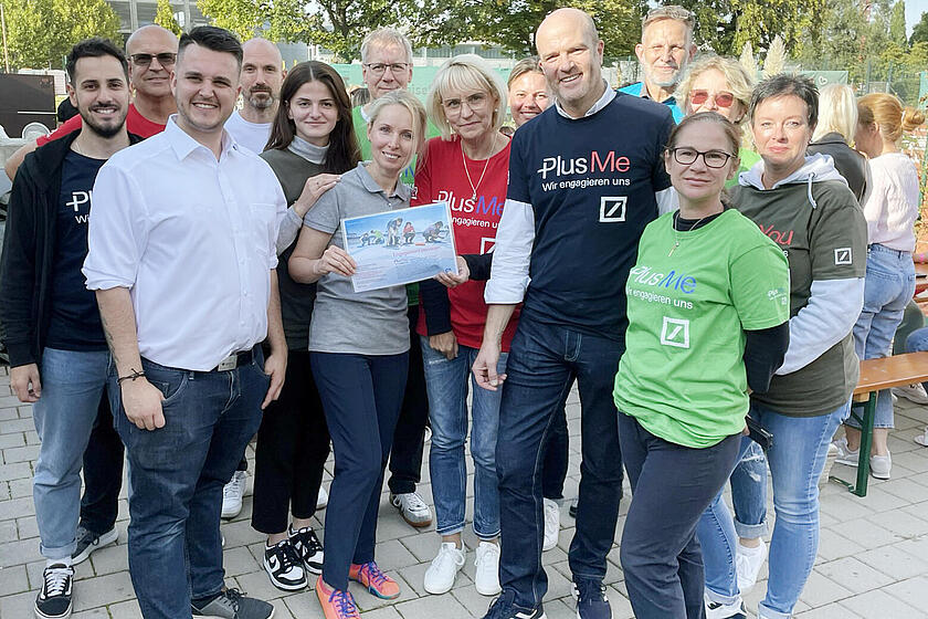 Eine große Gruppe an Leuten stehen auf einem Innenhof. Einige tragen ein T-Shirt mit der Aufschrift „Plus Me – wir engagieren uns“.