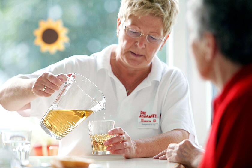 Eine ältere Frau schenkt einer weiteren Person an einem Tisch Trinken in ein Glas in einer hellen, freundlichen Umgebung.