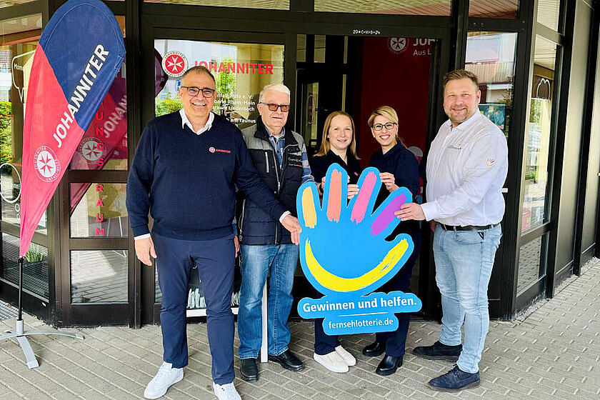 Fünf Personen stehen vor dem Gebäude der Johanniter-Unfall-Hilfe in Liederbach am Taunus und halten gemeinsam ein großes, farbenfrohes Hand-Symbol mit dem Schriftzug „Gewinnen und helfen – fernsehlotterie.de“.
