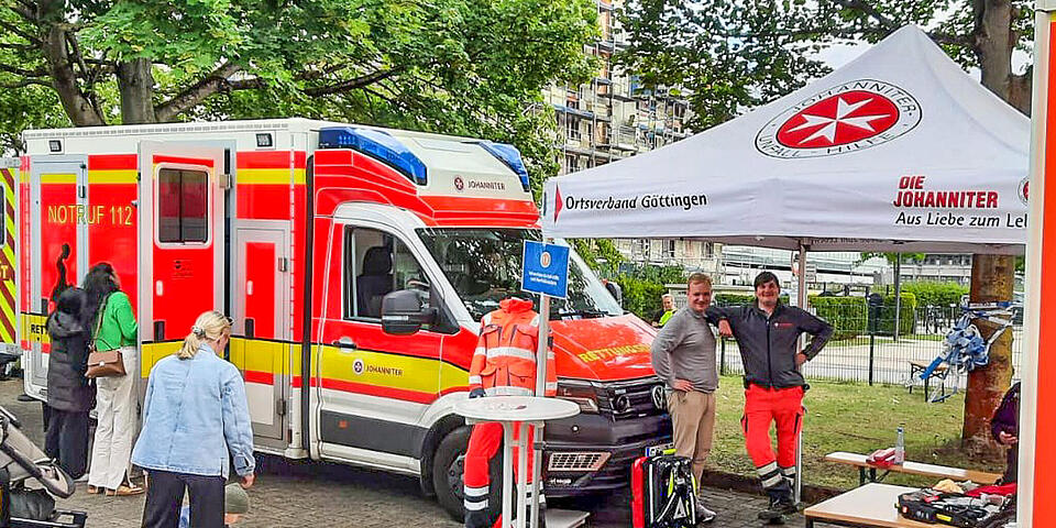 Zwei RTWs der Johanniter auf einem Parkplatz. Menschen schauen in einen der RTWs hinein. Im Hintergrund sind Bäume