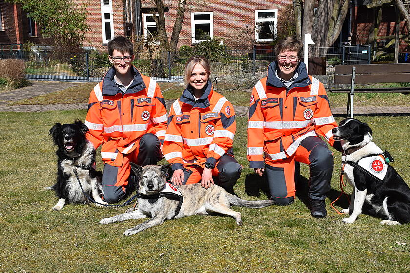 drei Hund-Mensch-Teams hocken auf dem Rasen und freuen sich über bestandene Prüfung